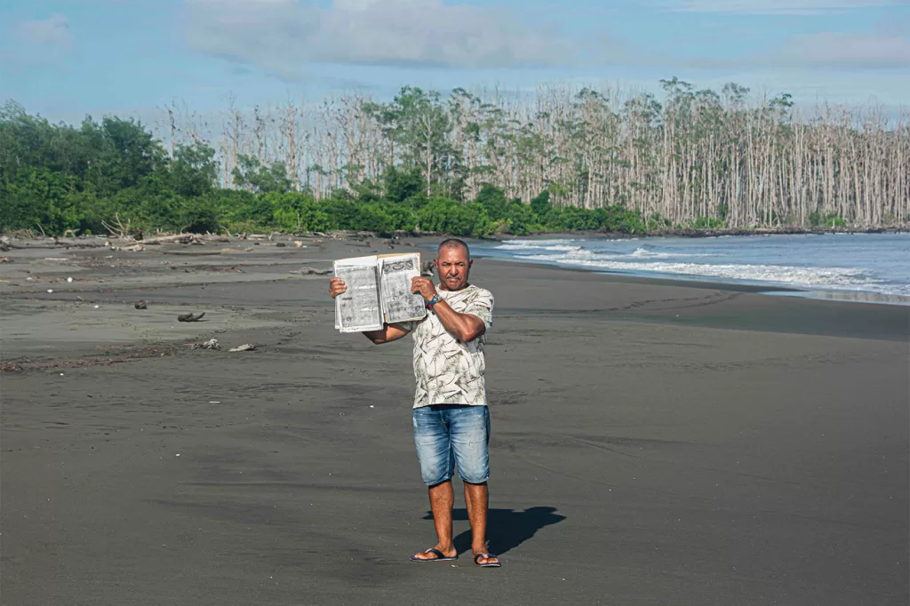 En la playa de Amarales, con Manglares deshojados de fondo, el representante legal del Consejo Comunitario Punta Mulatos, Robinson Campaz, sostiene una copia de la escritura de la propiedad colectiva de las playas hoy afectadas por la erosión, que data de finales del siglo XVIII, cuando el territorio era más grande. Fotografía: Manuel José Cárdenas Martínez.